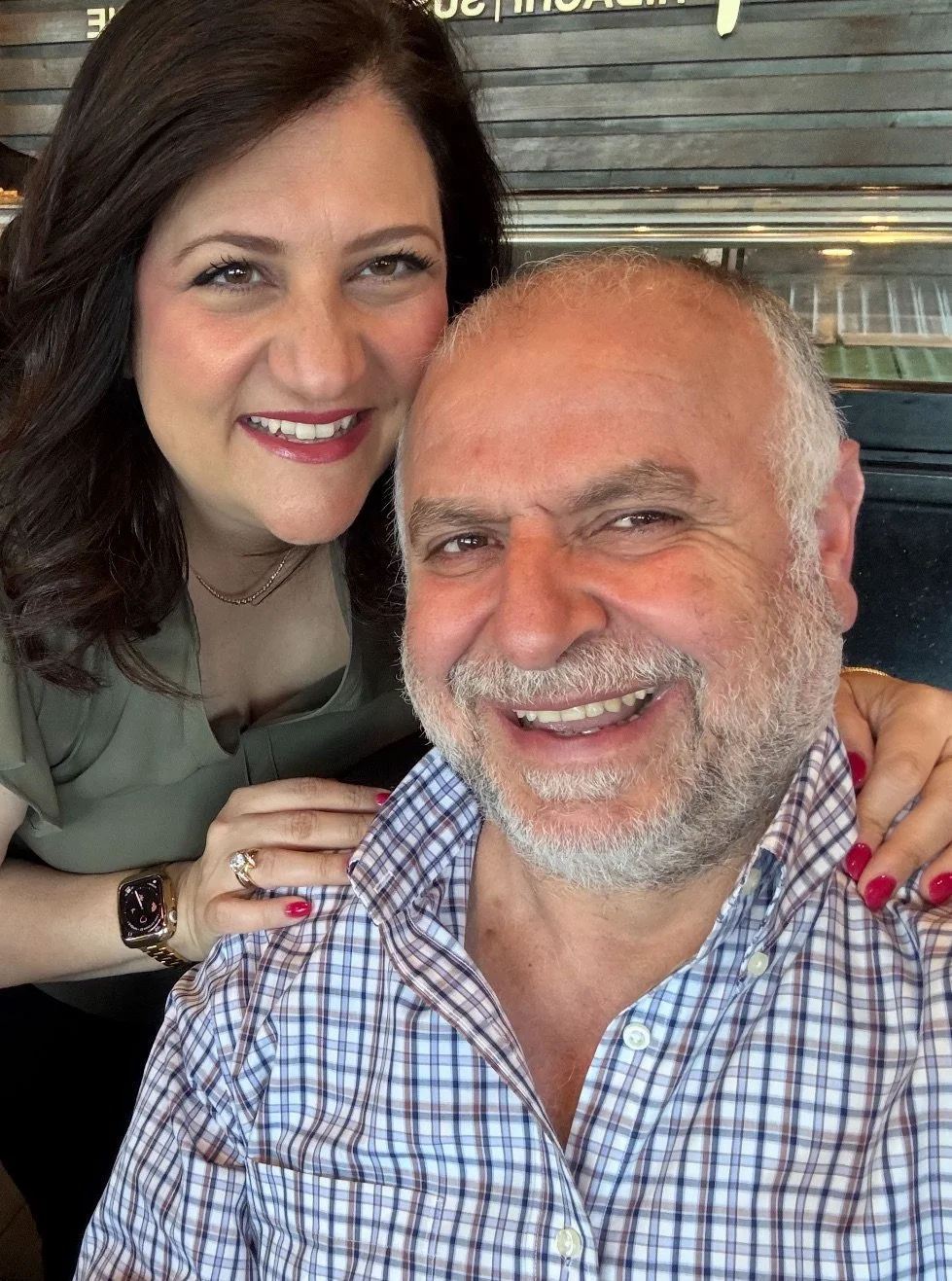 A woman with dark hair smiling and a man with gray hair and beard smiling, taking a close-up selfie together.