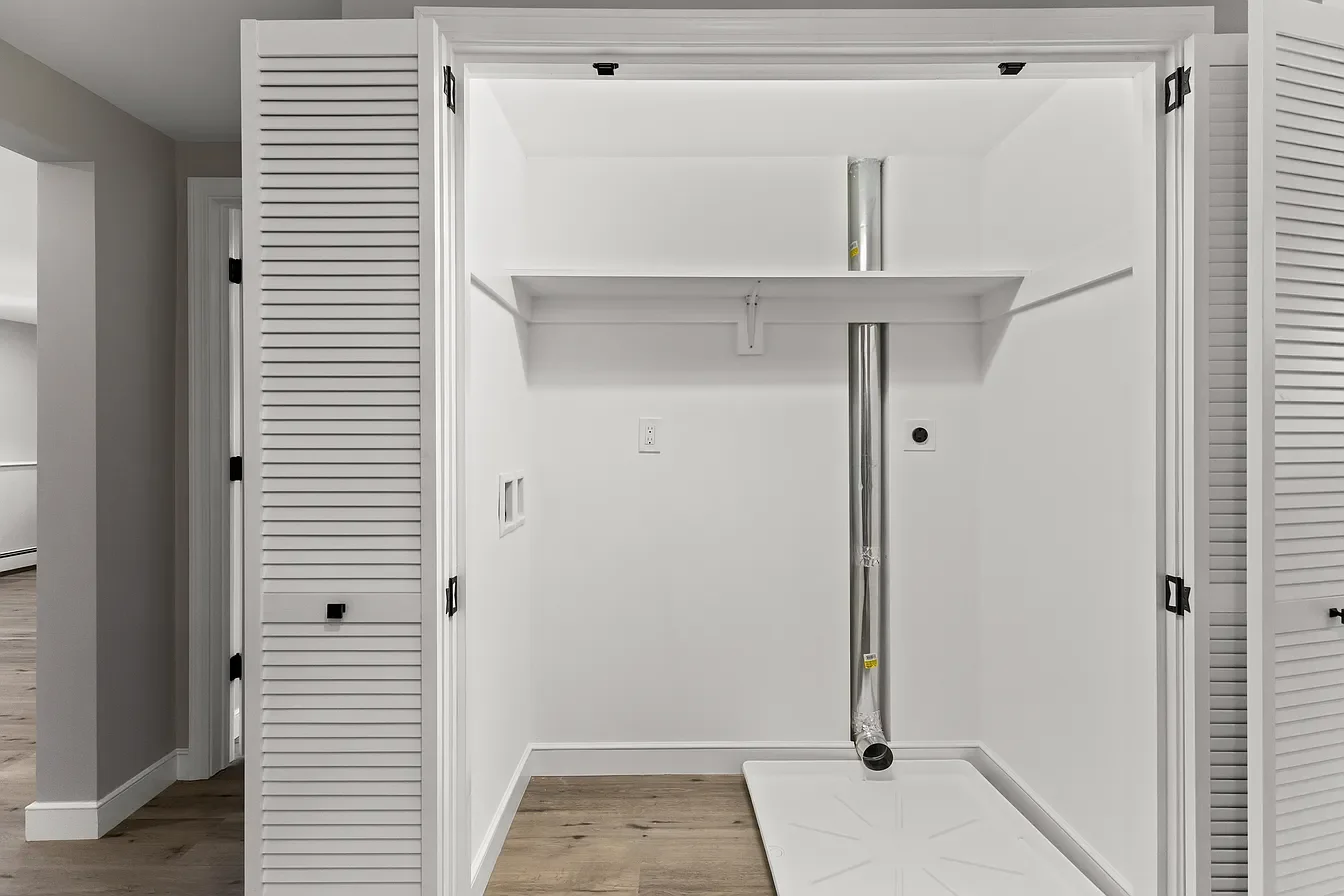 Empty laundry room with white walls, louvered closet doors, and ventilation pipe, with brackets for a washer and dryer and a drain pan on the floor.