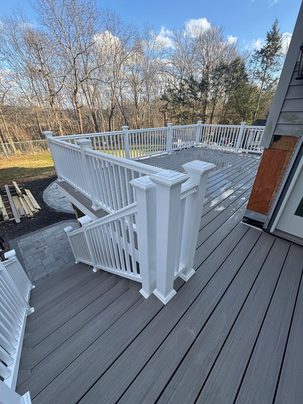 Newly built wooden deck with white railing attached to a house, overlooking a backyard with trees and a cloudy blue sky.