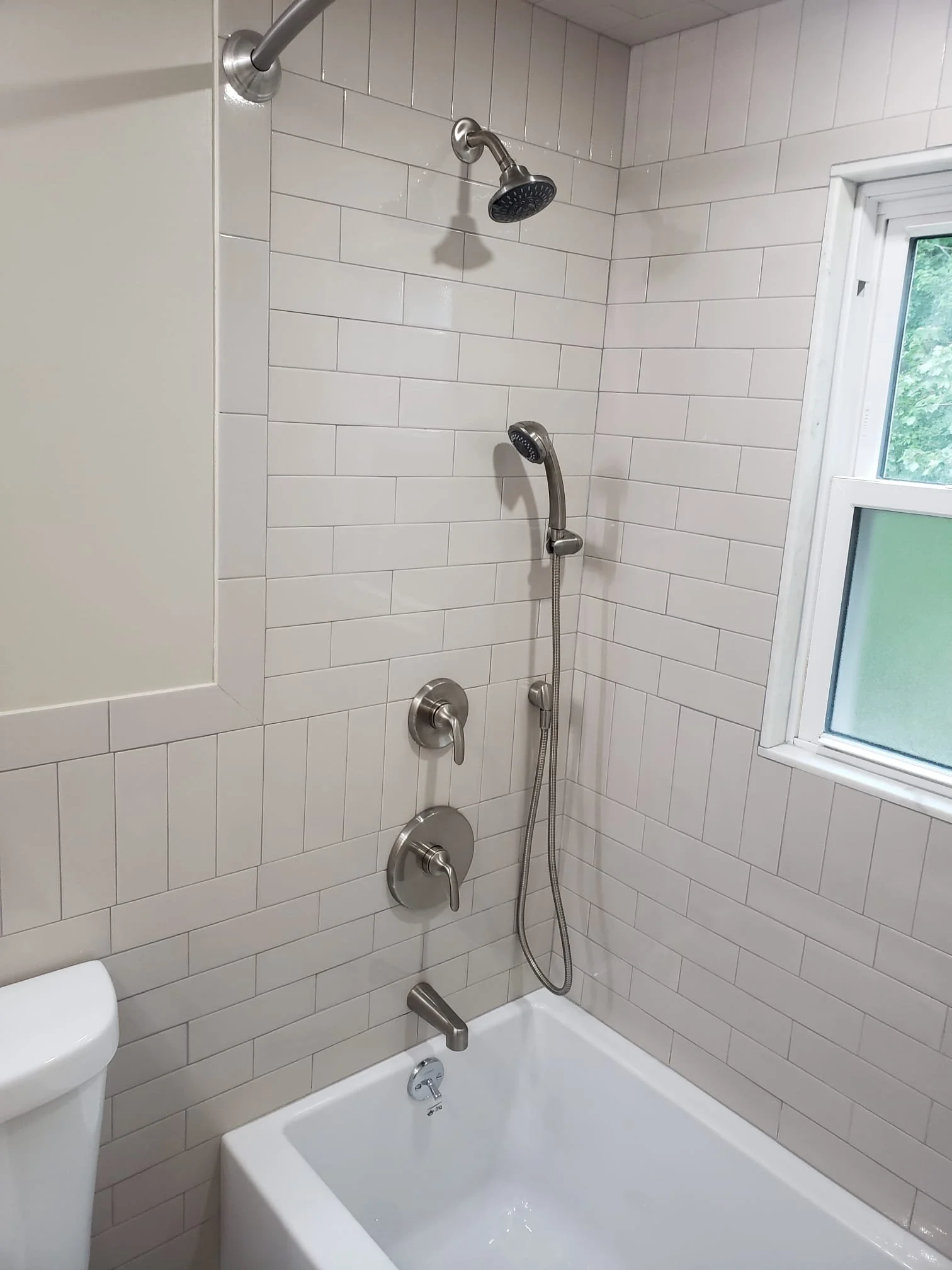 Bathroom shower with a handheld and fixed showerhead, white subway tile walls, a window, and a bathtub.