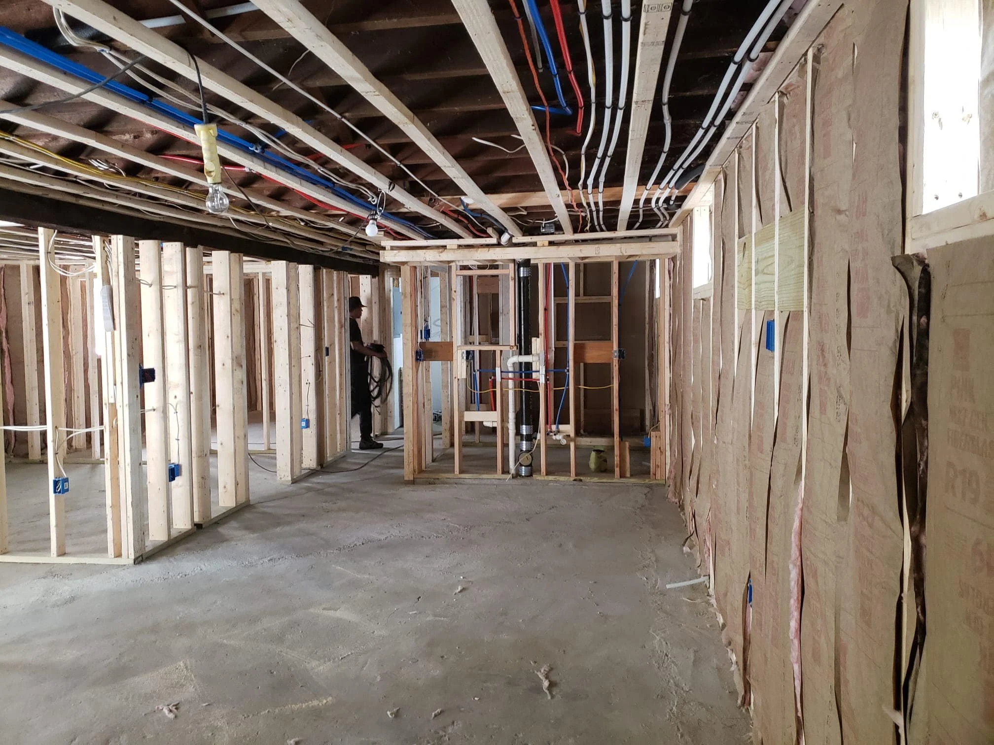 Interior view of a house under construction, showing exposed wooden wall framing, ceiling with electrical wiring, and a worker in the background.
