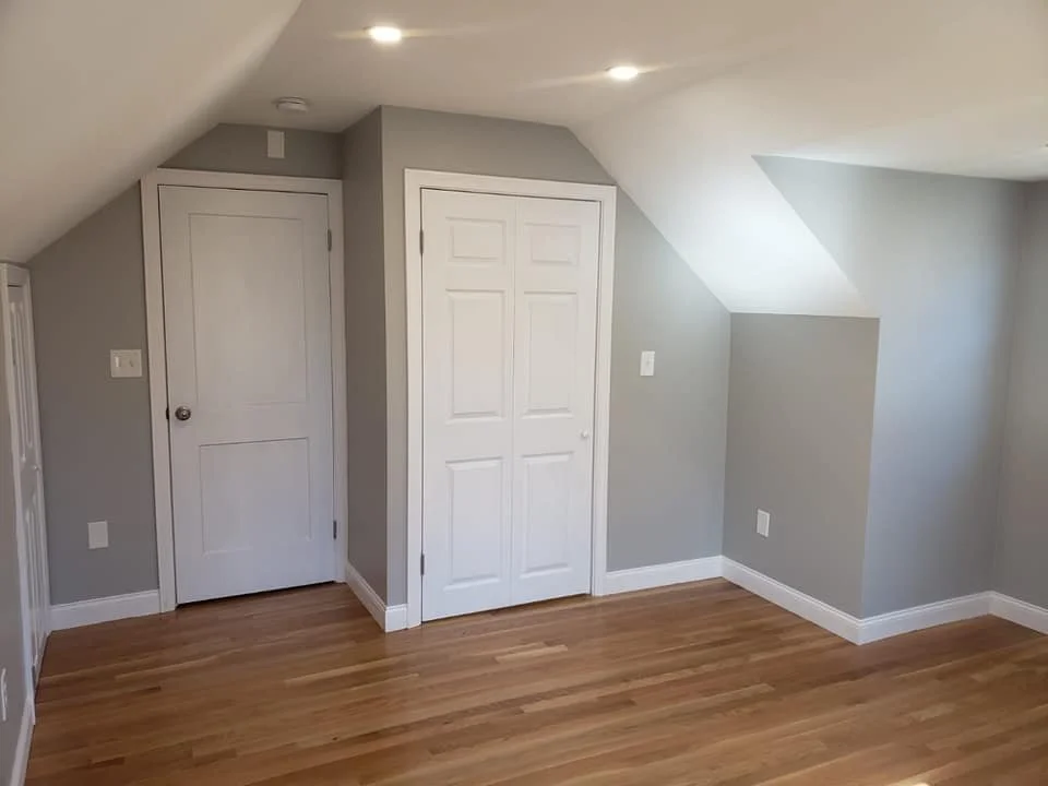 Empty room with hardwood floors, gray walls, a small closet with a folding door, a regular door, and a sloped ceiling with recessed lighting.
