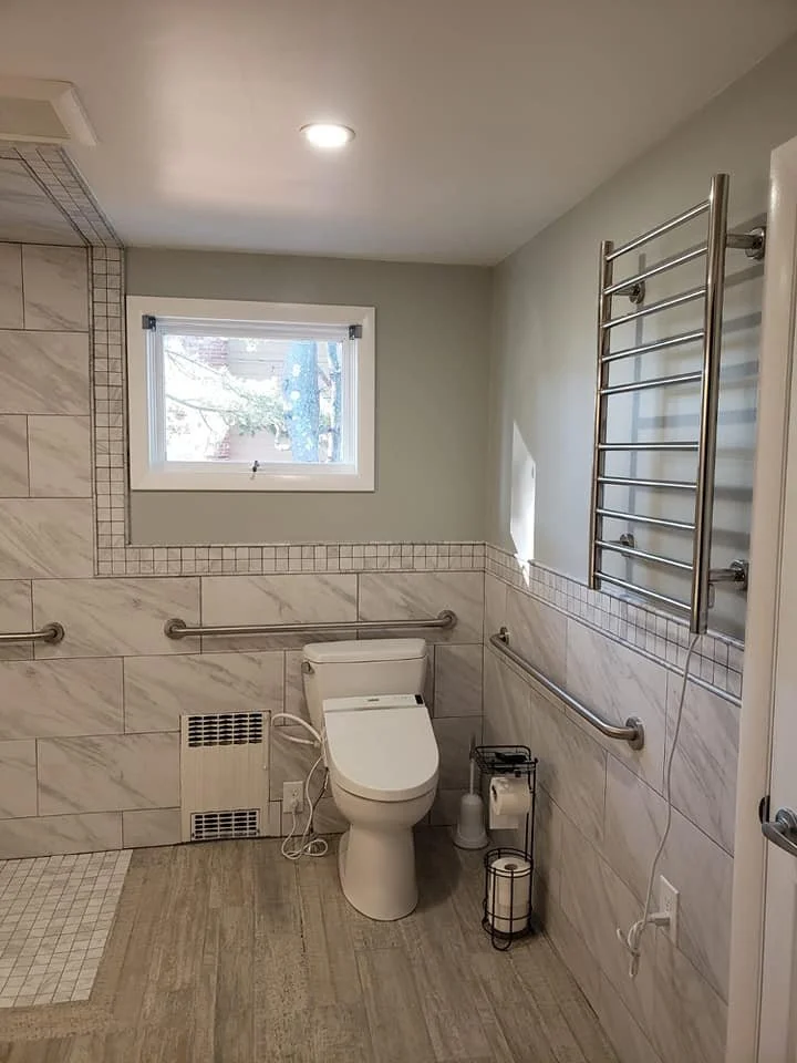 Accessible bathroom with a toilet, a window, white tile walls, wood floor, and metal grab bars.