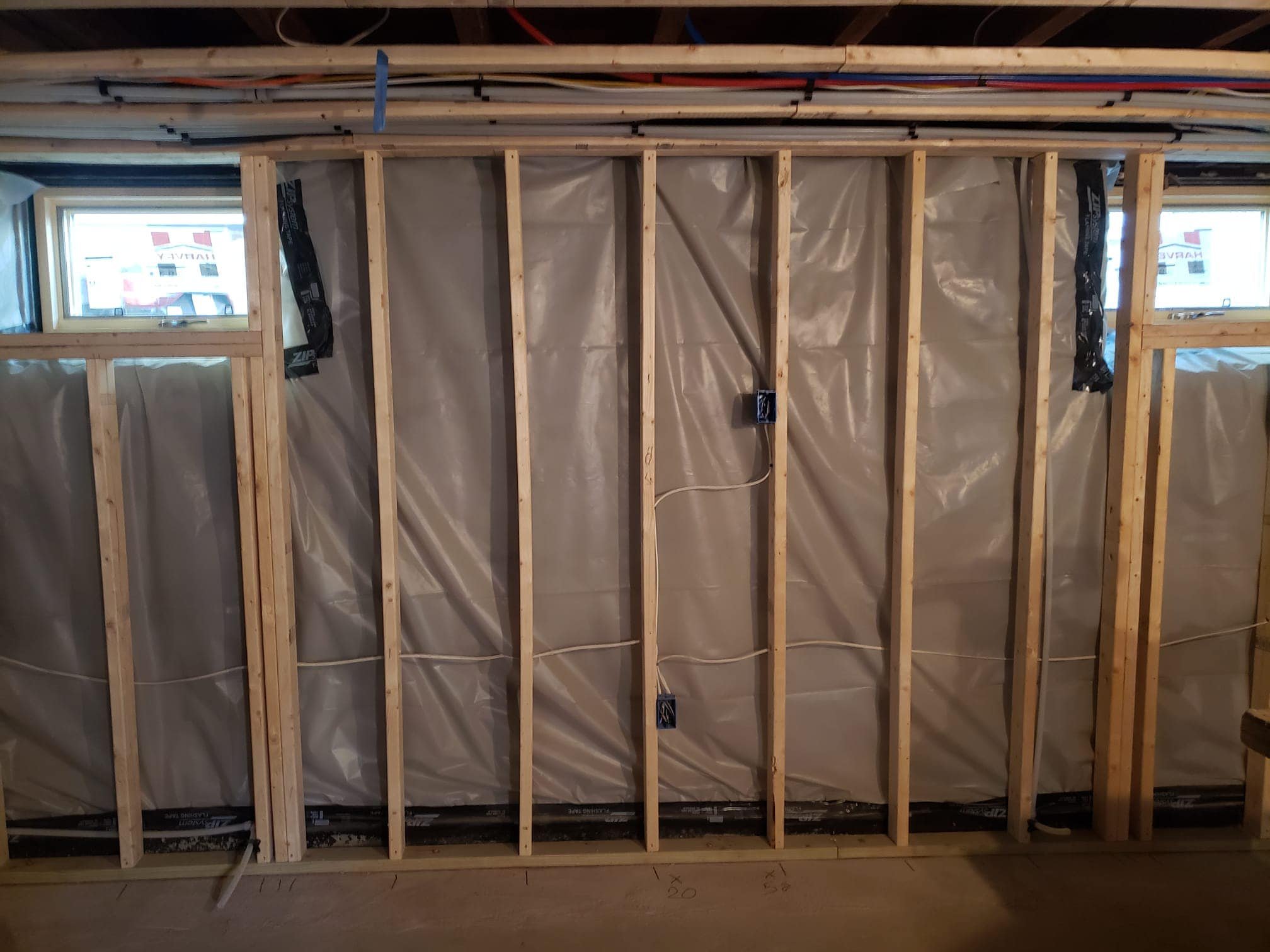 Interior of a building under construction with visible wooden framing, plastic vapor barrier, electrical wiring, and small rectangular windows in the walls.