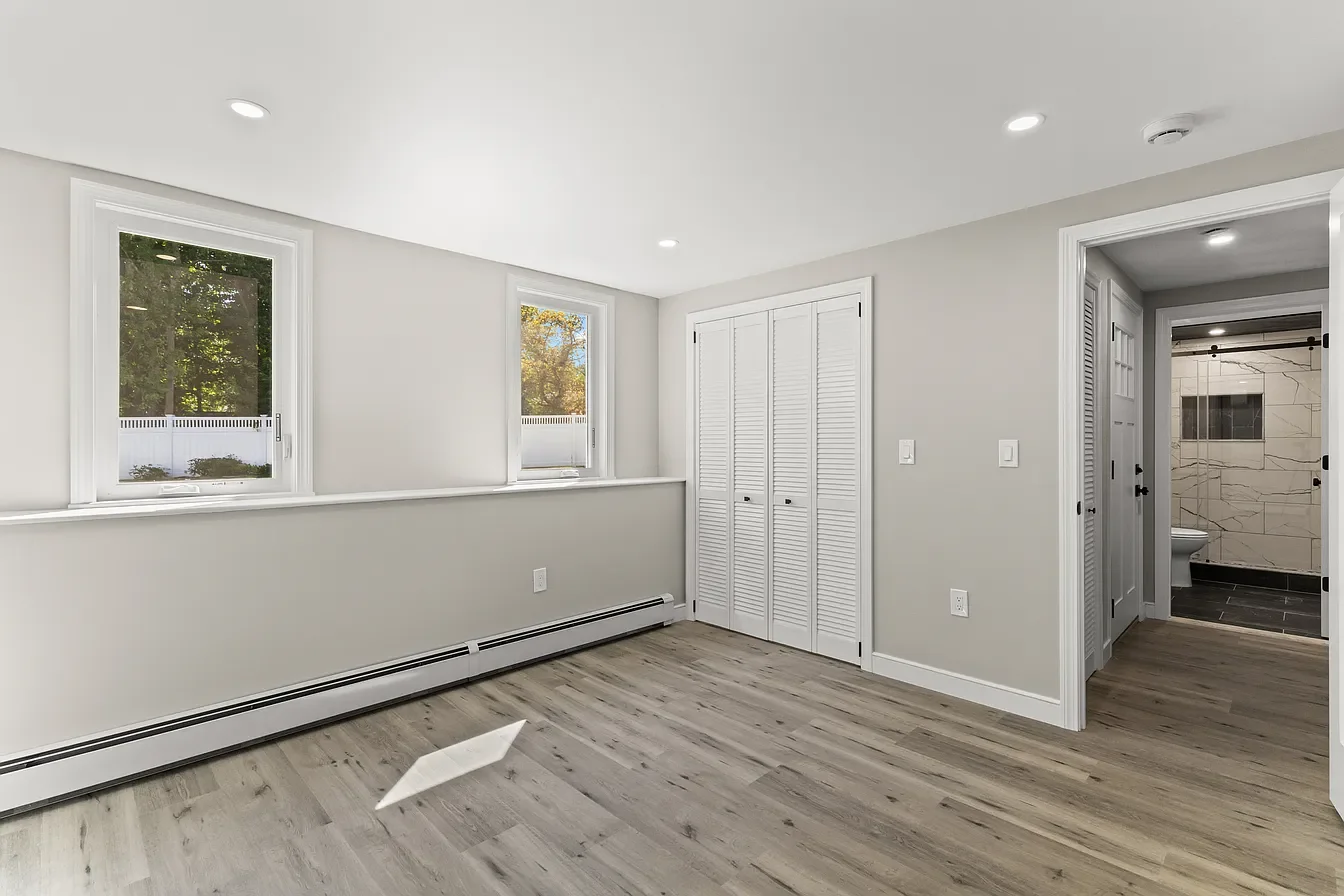 Empty room with light gray walls, wood flooring, two windows, open closet with louvered doors, and a bathroom with marble tiled walls in the background.