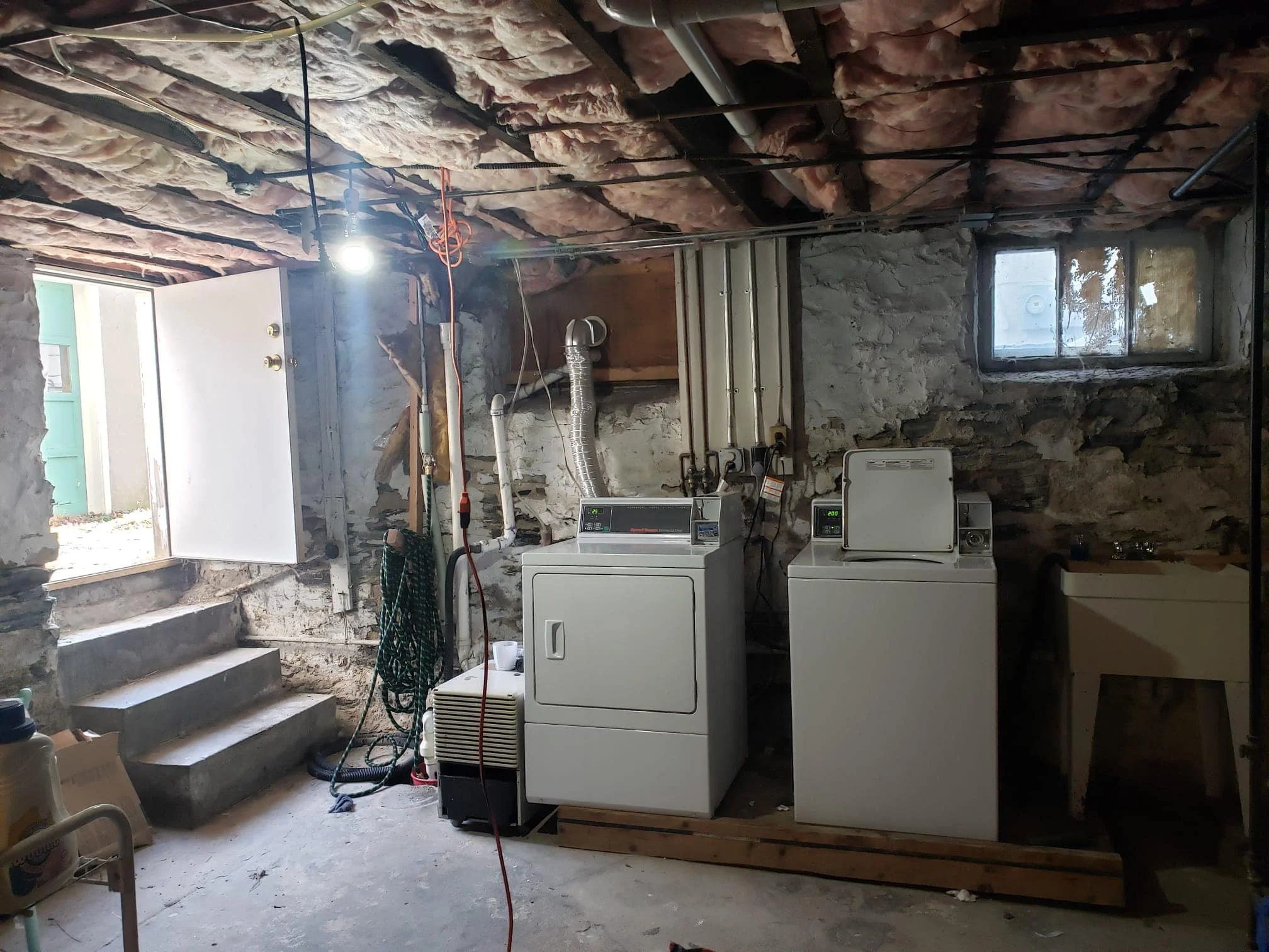 Laundry room with washing machine and dryer, stone walls, small window, open door, concrete stairs leading outside, and miscellaneous items.