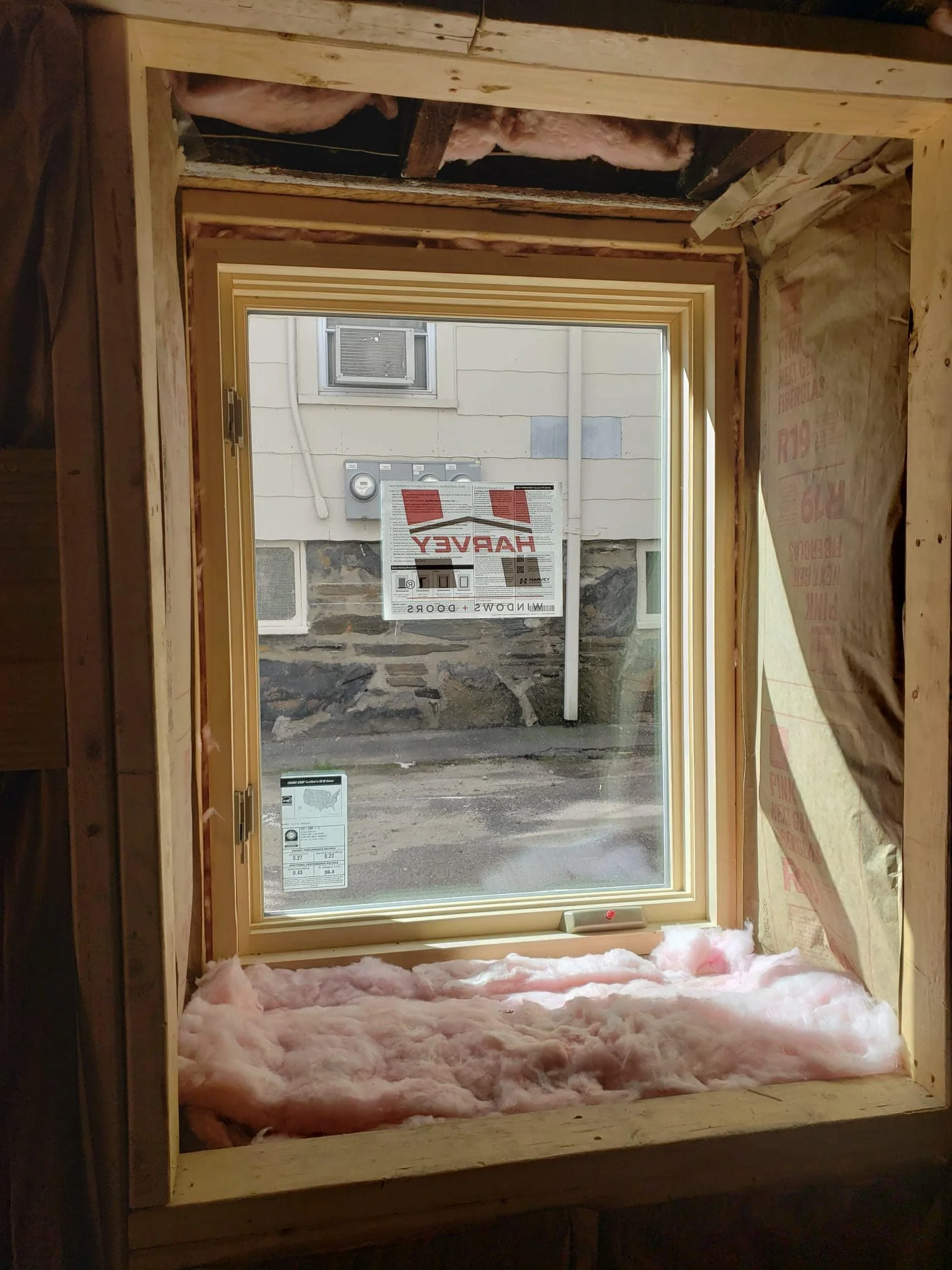 View of a new window with installed insulation surrounding it, showing pink fiberglass insulation at the bottom and partially on the sides, with an outside view of a stone wall and window.