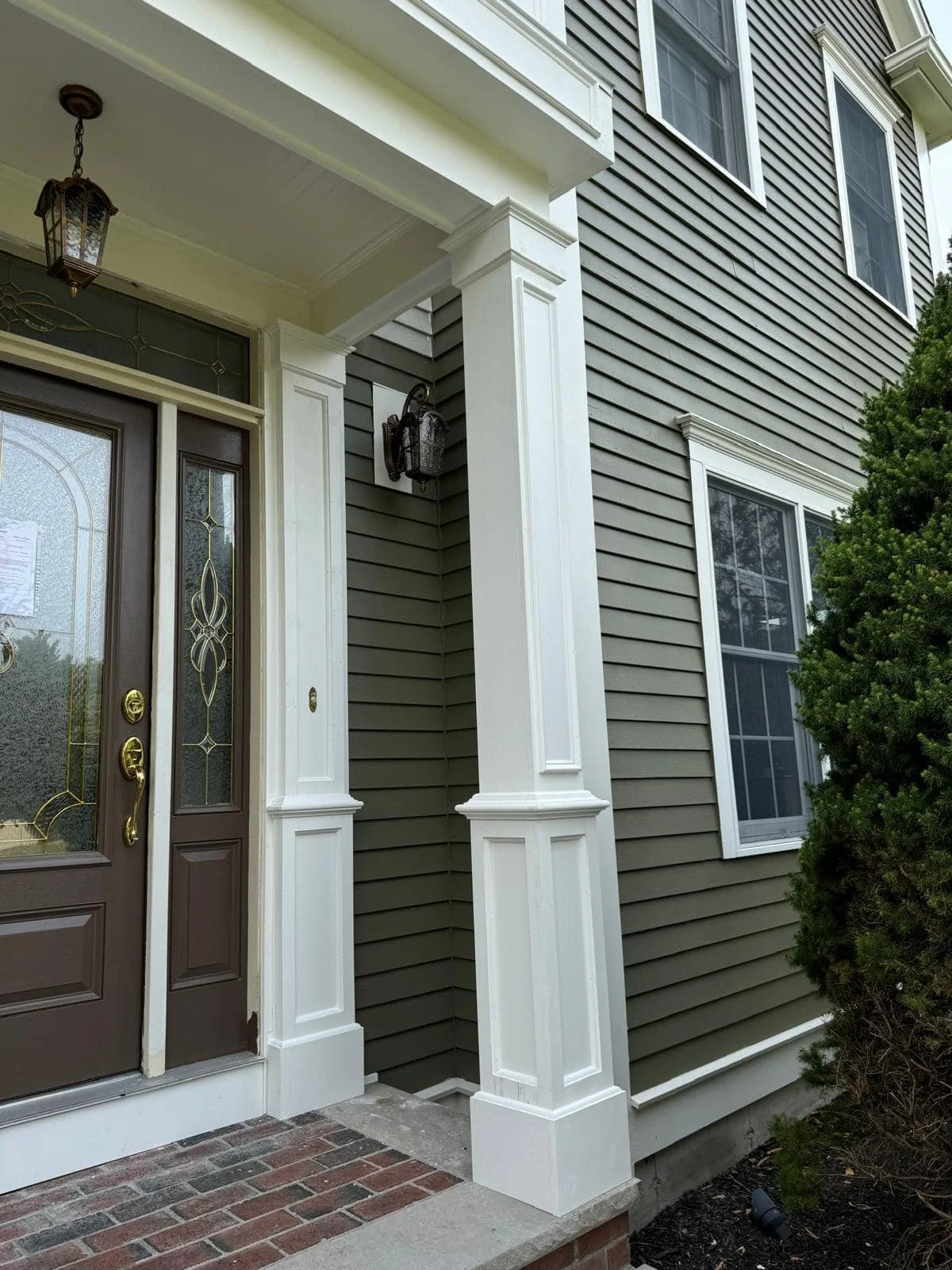 Close-up of the front porch of a house with a dark brown door, decorative glass panels, white trim, and black lantern-style light fixtures, with brick steps and a side garden with bushes.
