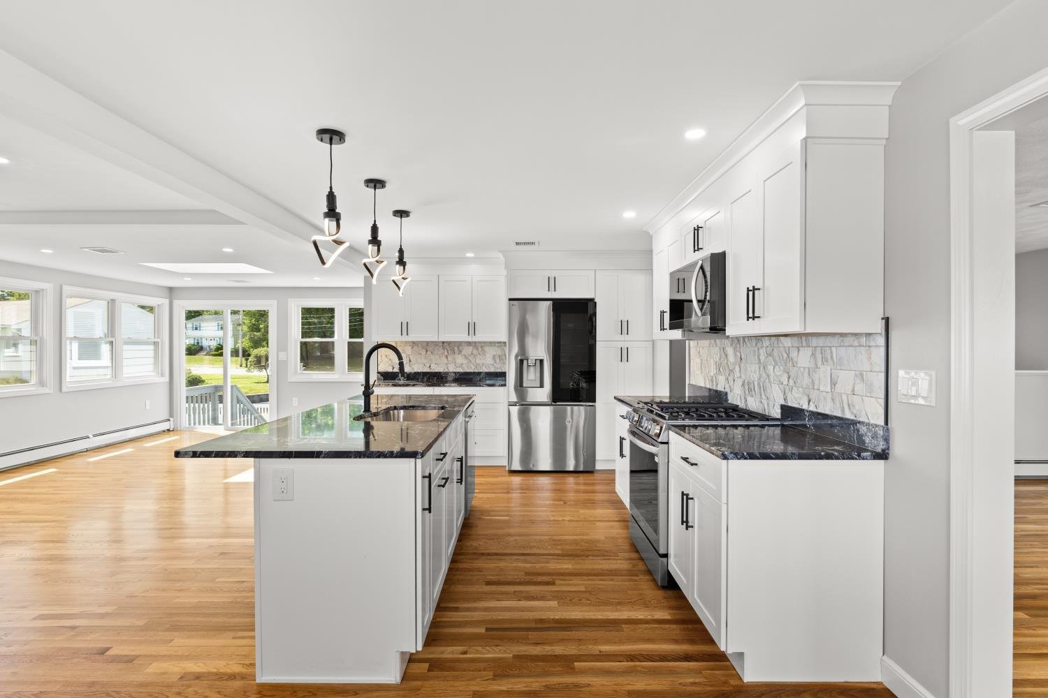 Modern kitchen with white cabinets, black countertops, stainless steel appliances, and hardwood floors, featuring a central island and large windows for natural light.