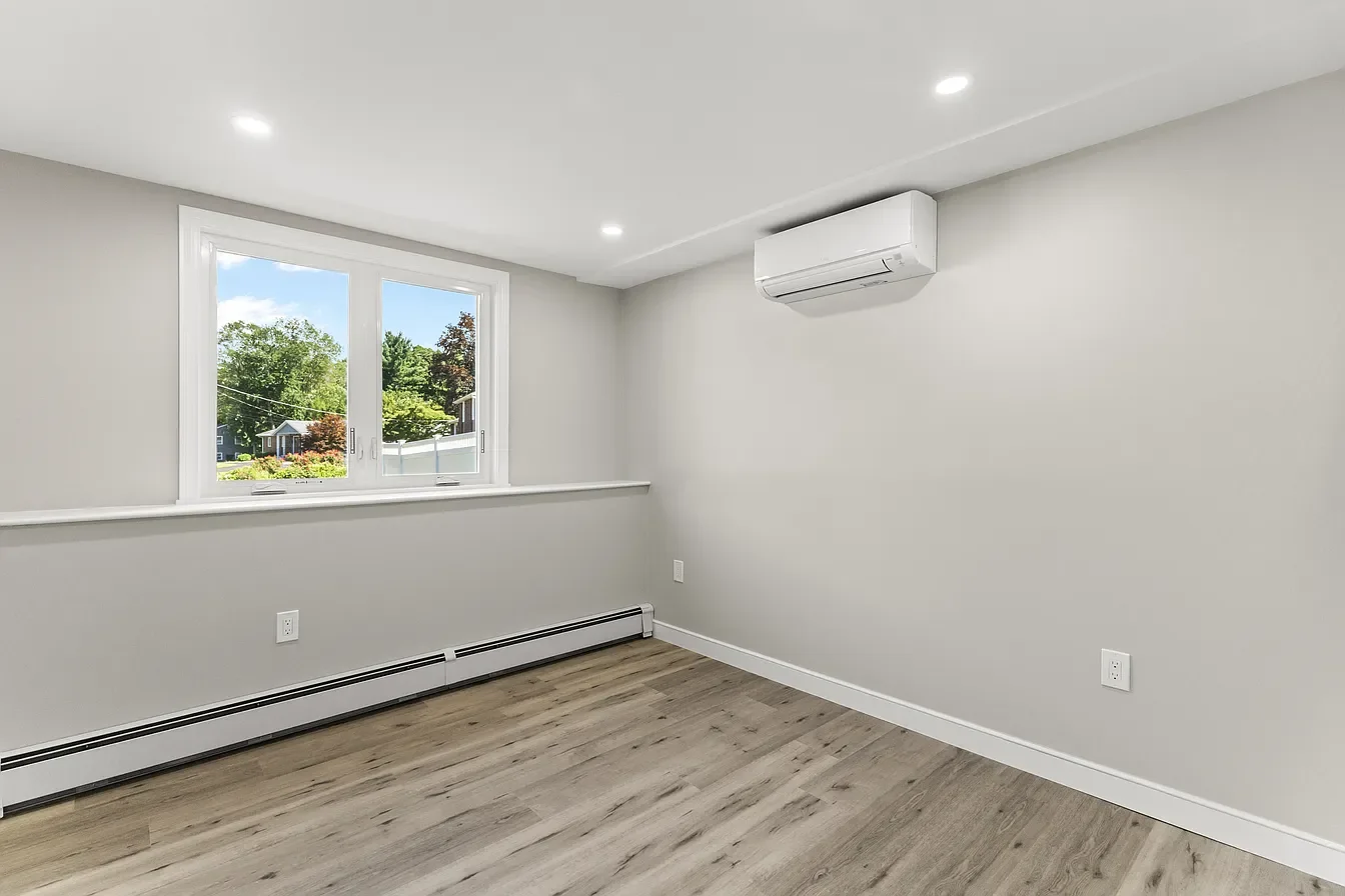 Empty room with white walls, hardwood floor, a window with a view of trees and houses outside, a wall-mounted air conditioner, and ceiling lights.