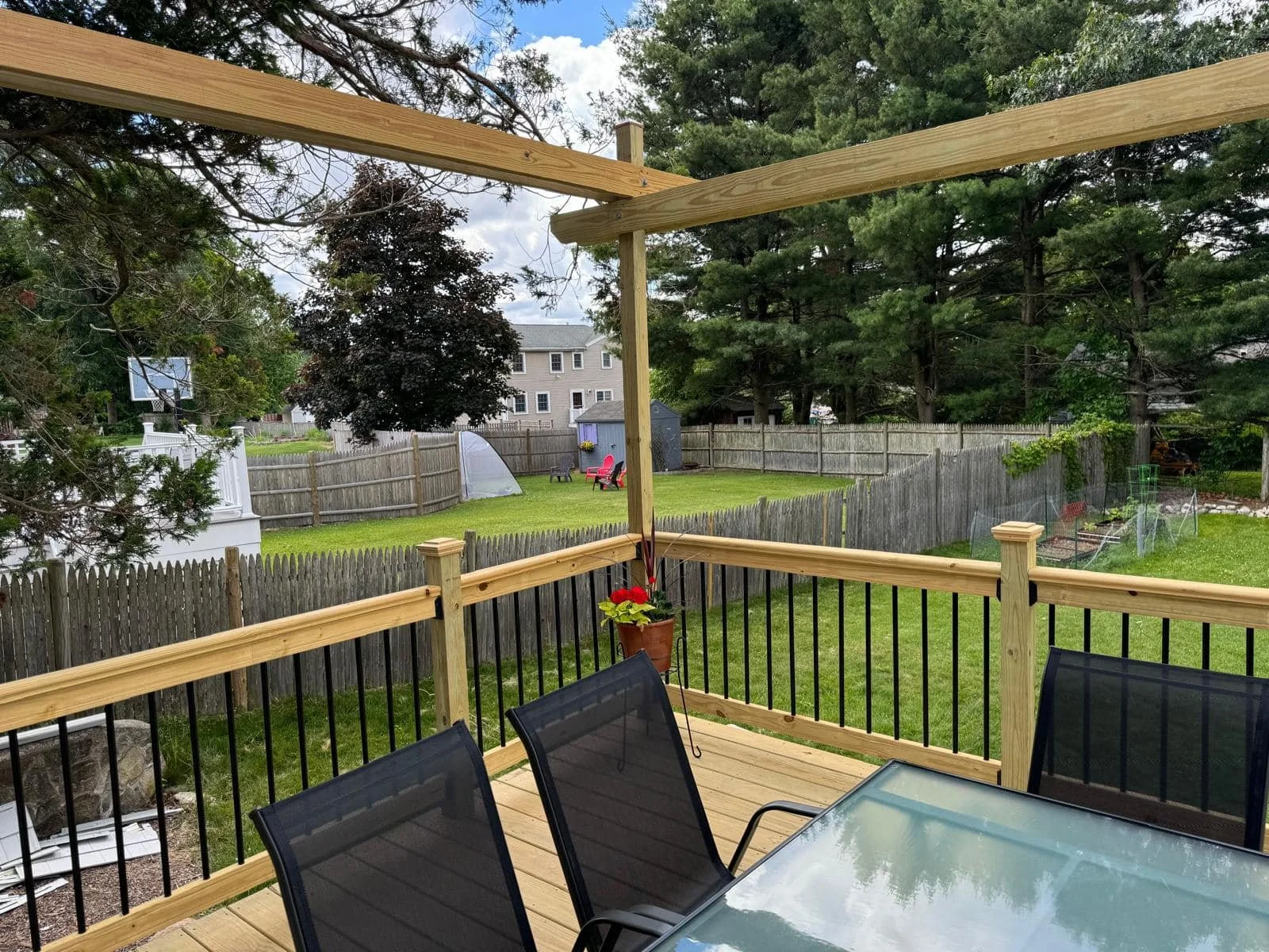 A wooden deck with a glass-top table and black chairs overlooking a backyard with green grass, trees, and a wooden fence.