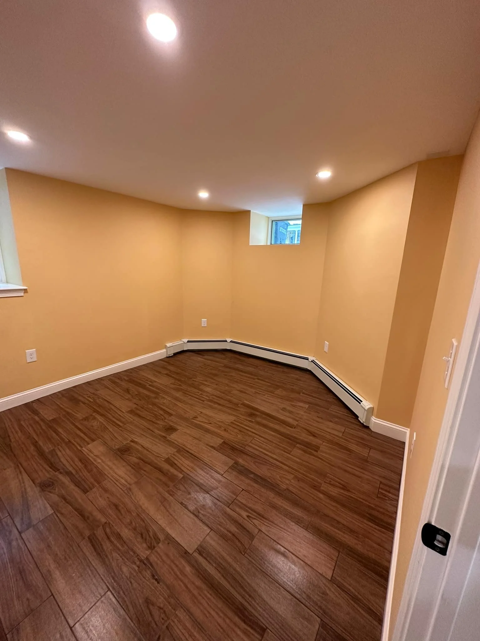 Empty room with beige walls, wood floor, small window, ceiling lights, and baseboard heater