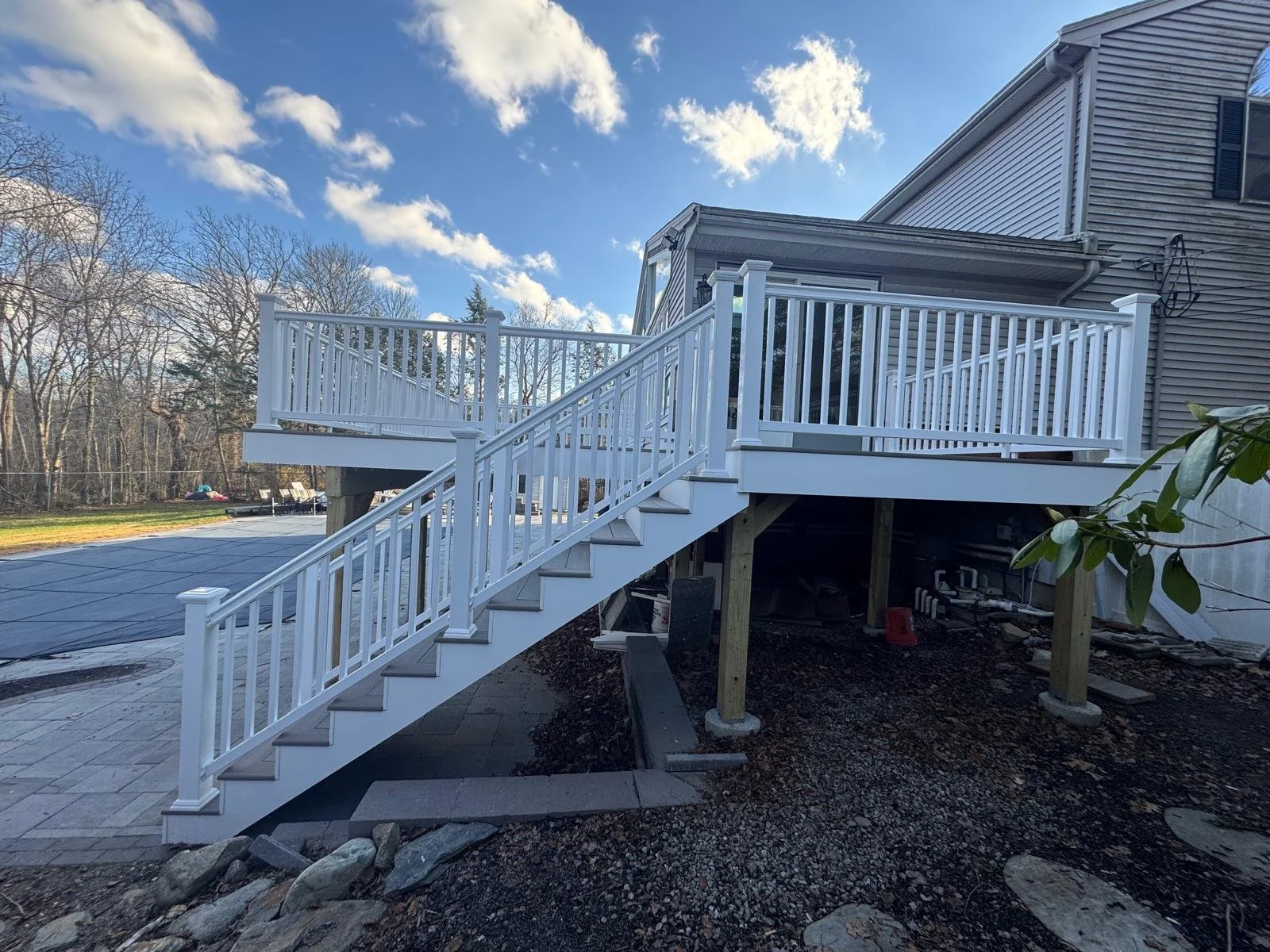 Newly constructed white wooden deck with stairs outside a house, supported by wooden posts, with trees and a blue sky in the background.