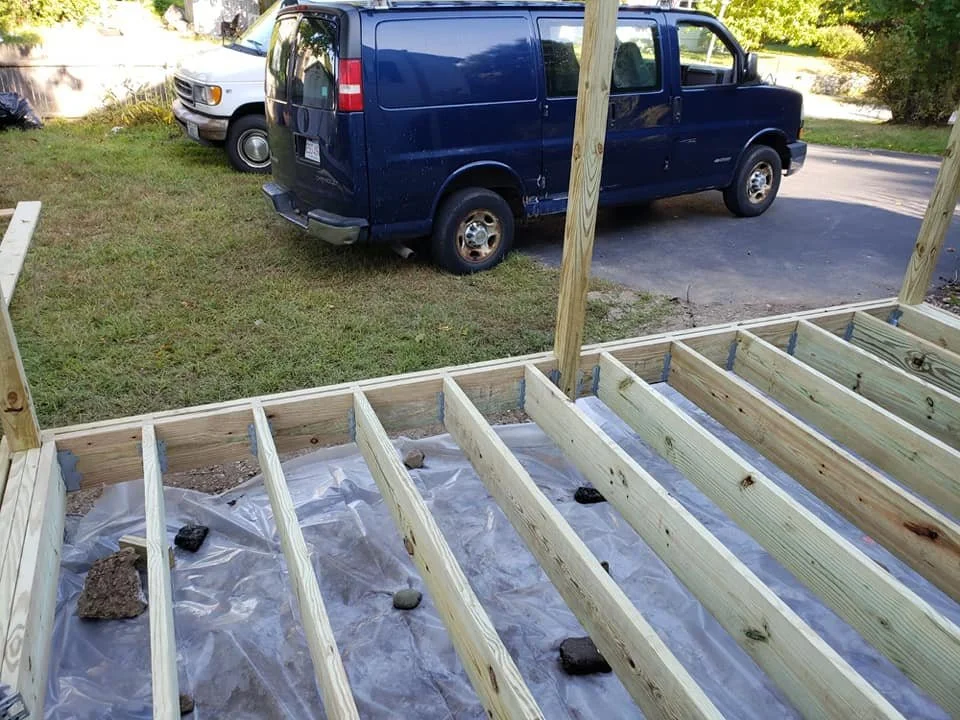 Construction site with wooden framing for a deck or patio, placed on plastic sheeting with rocks for weight, next to parked vehicles including a blue van and an older white minivan, on a grassy area with trees and street.