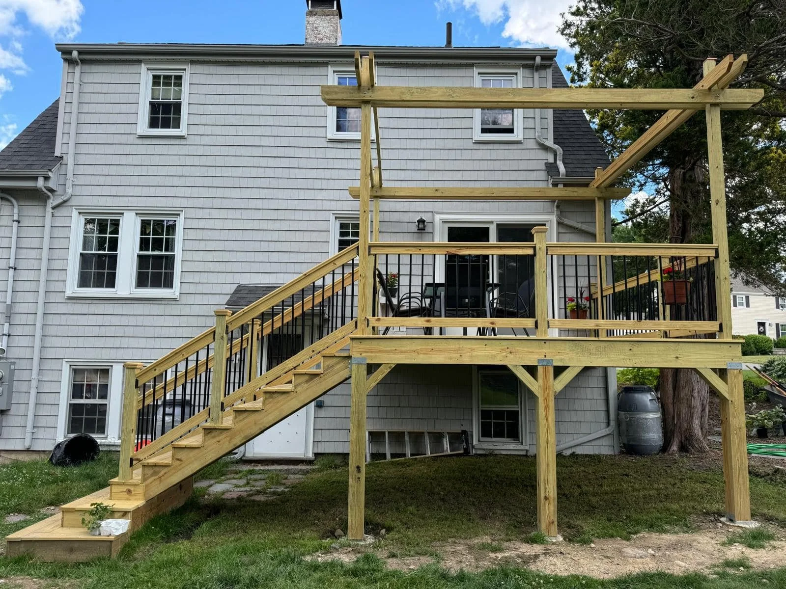 Newly built wooden deck with stairs and railing attached to the back of a two-story house with gray siding, two-stall windows, and sliding glass door; outdoor furniture and potted plants on the deck; large tree to the right.