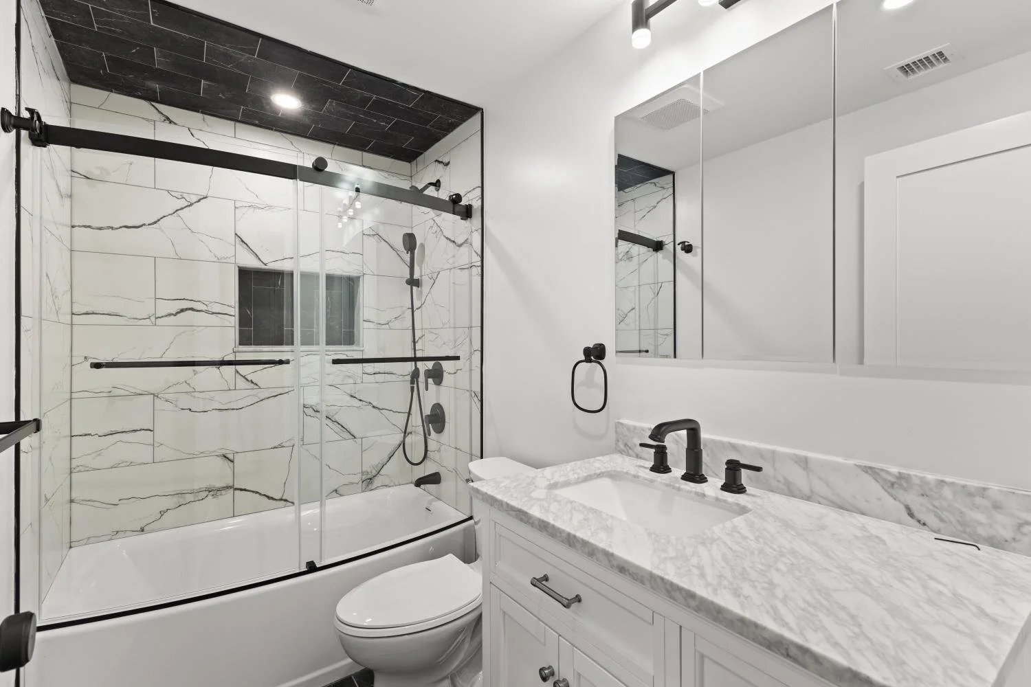 Modern bathroom with white vanity, black fixtures, marble countertop, and a walk-in shower with glass doors and black hardware, marble wall tiles, and a ceiling light.