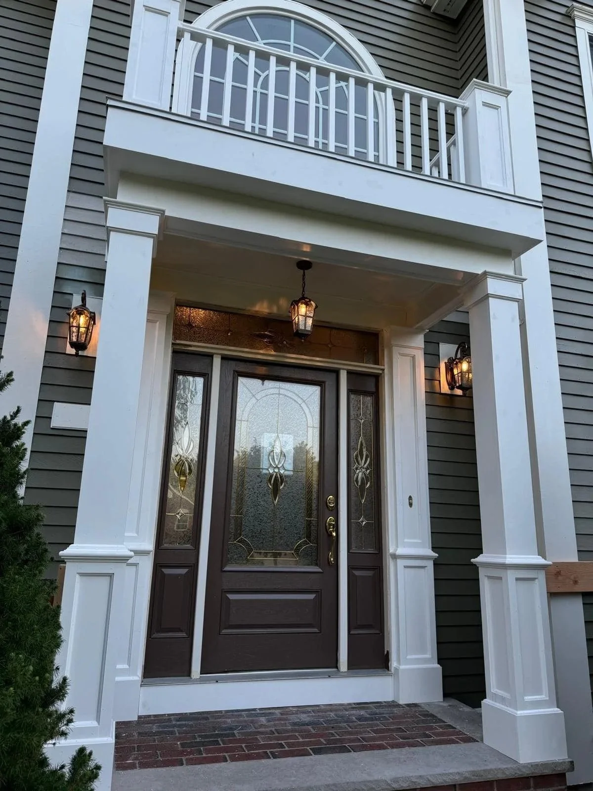 Front porch of a house with a dark wooden door featuring decorative glass panels, white framing, and exterior lighting fixtures on either side of the entrance. There is a balcony with a white railing above.