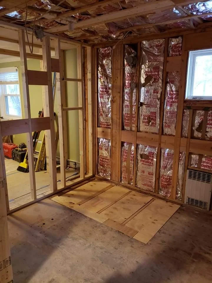Interior of a house under construction with exposed wall studs, insulation, and a partially finished floor.