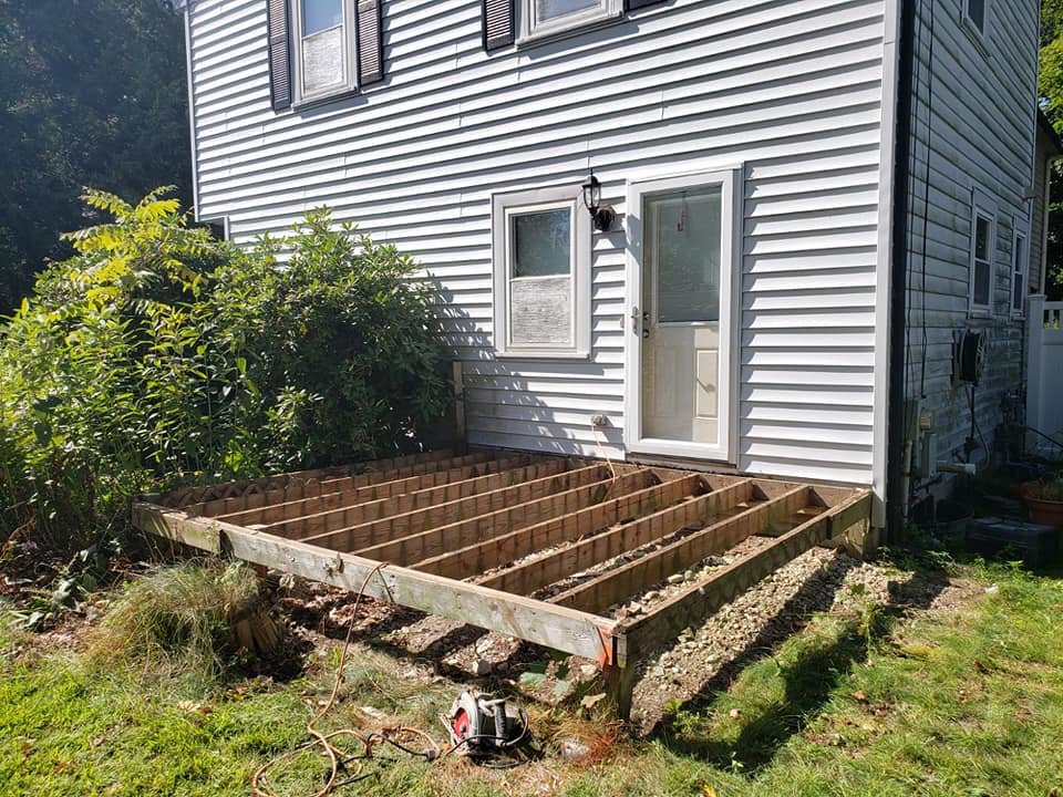 Construction of a wooden deck in progress outside a house with white siding, a door, and windows. Construction tools and materials are present, with a garden to the side.