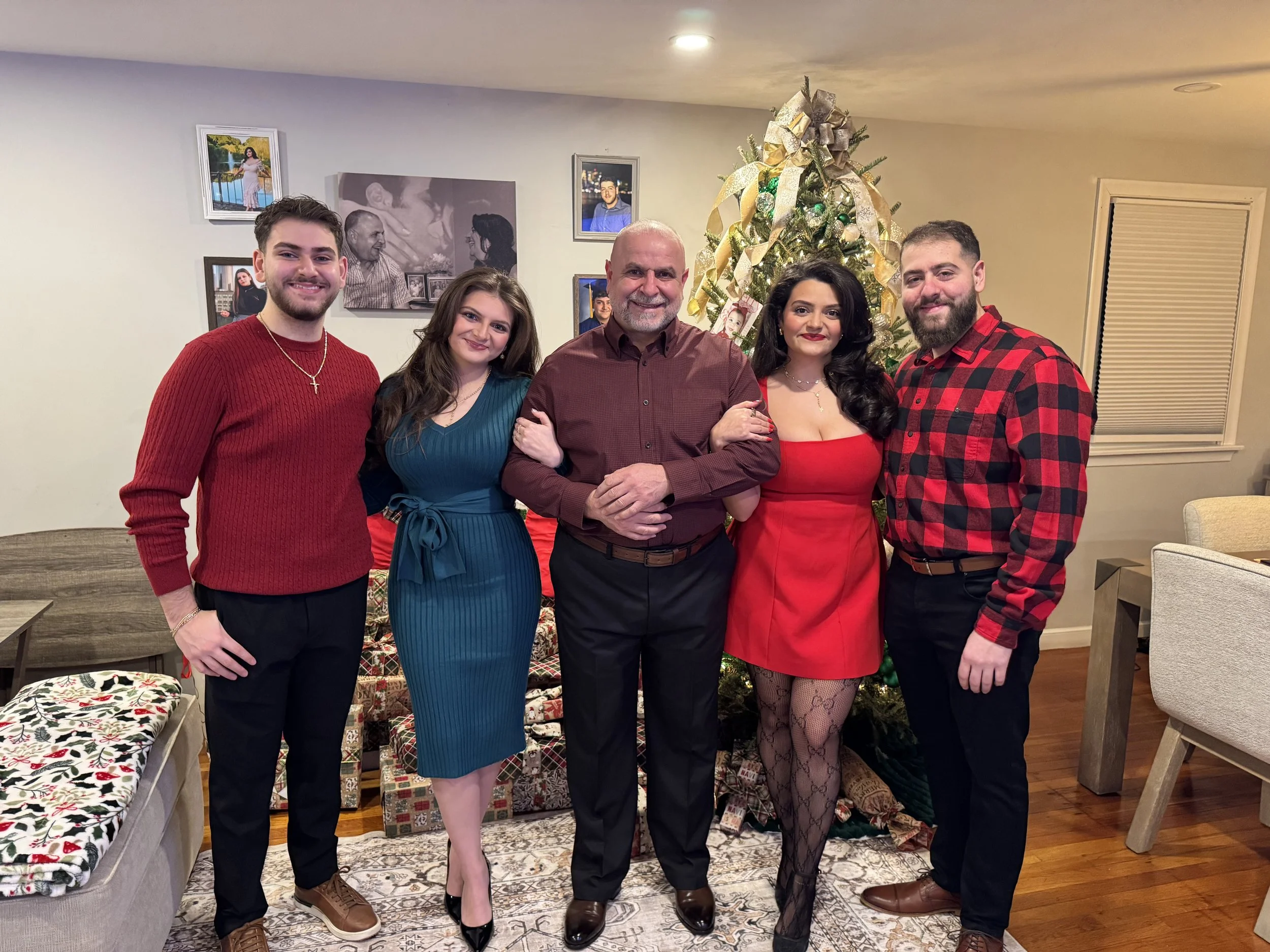 A family of five standing together in a living room decorated for Christmas with a Christmas tree and wrapped presents. The family includes two men, two women, and a man with a mustache and beard. They are all dressed in holiday attire, smiling, with the Christmas tree in the background.