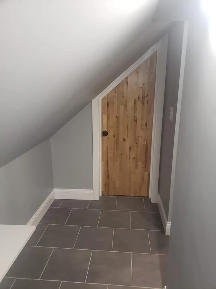 A small, sloped ceiling room with gray tiled floor, white baseboards, and light gray walls. There is a small wooden door on the sloped wall.