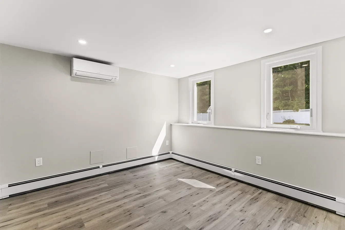 Empty white room with two windows, wood flooring, and an air conditioning unit on the wall.