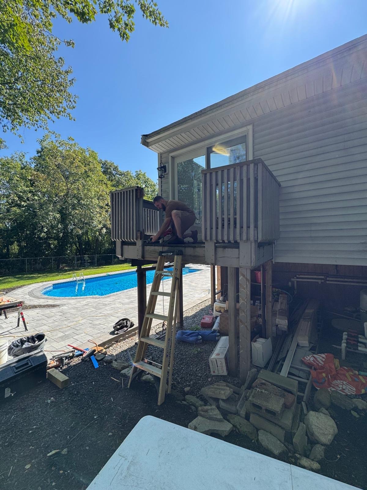 A man working on a small porch or deck attached to a house, with a backyard and swimming pool visible in the background, under a clear blue sky.