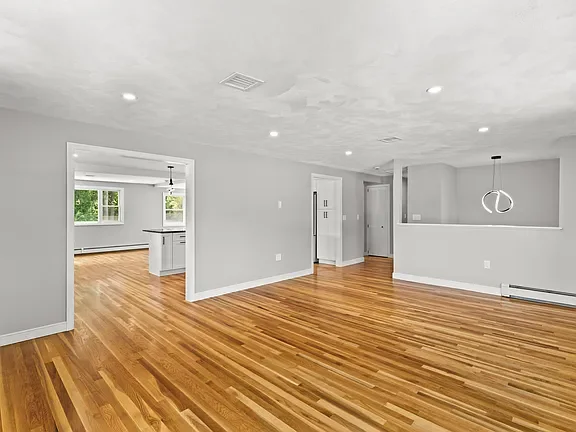 Empty living room with hardwood floors and white walls, connected to a kitchen area in the background.