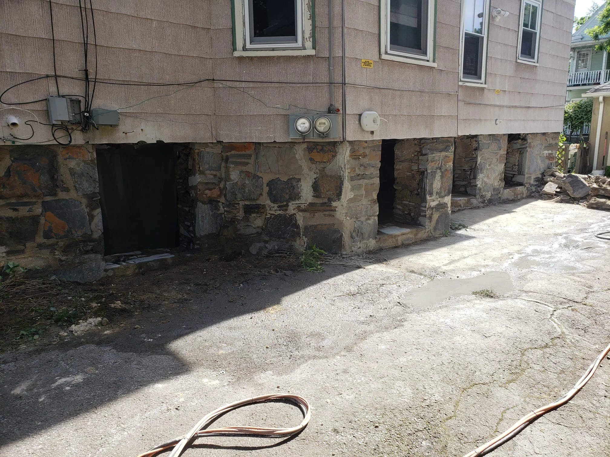 Basement of a house with stone foundation and small doorways, some blocked, with electrical meters and wires on the wall. Paved yard with cracks and a garden hose on the ground.