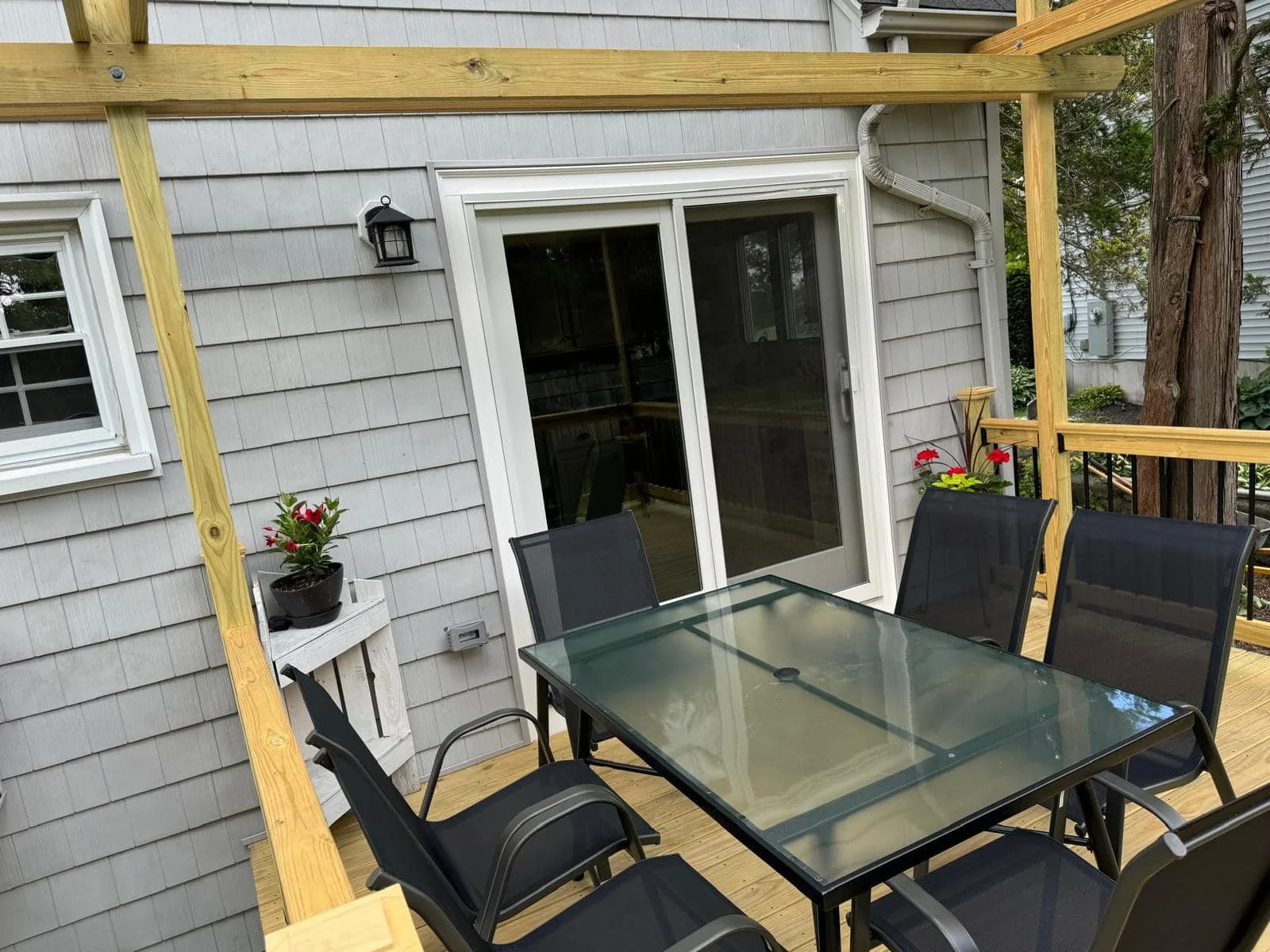 Outdoor deck with a glass-top table and six black chairs, attached to a house with gray siding and sliding glass door. There are potted red flowers on both sides of the door and a black wall-mounted lantern near the door.