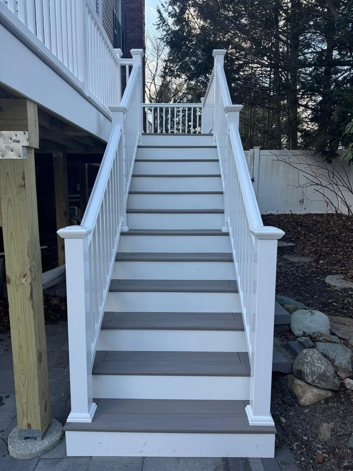 White outdoor staircase with grey steps and matching white railing, leading up to a deck on a house, with trees and a white fence in the background.