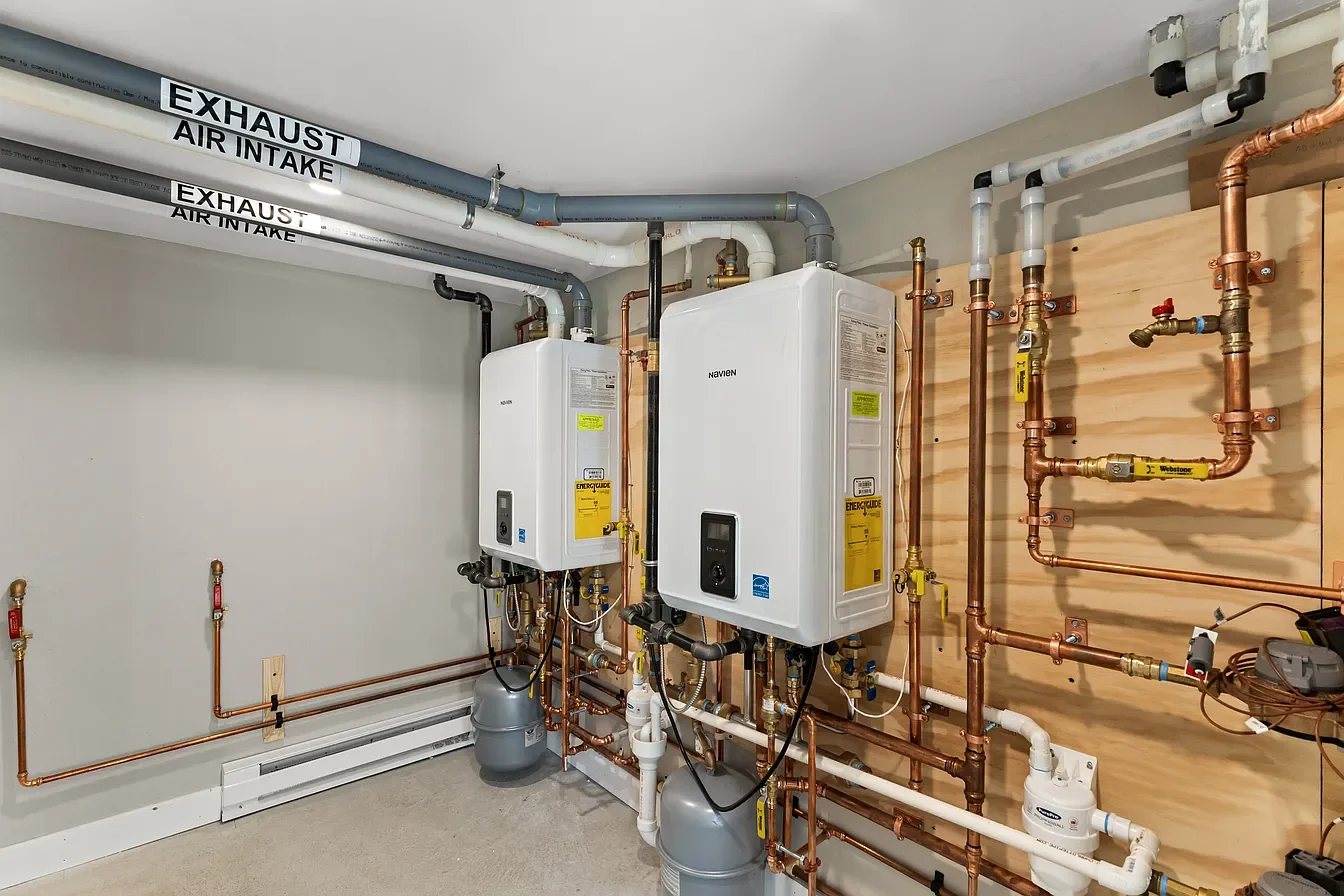 Indoor utility room with plumbing and heating system, including two water heaters, copper pipes, and labels for exhaust air intake and exhaust.