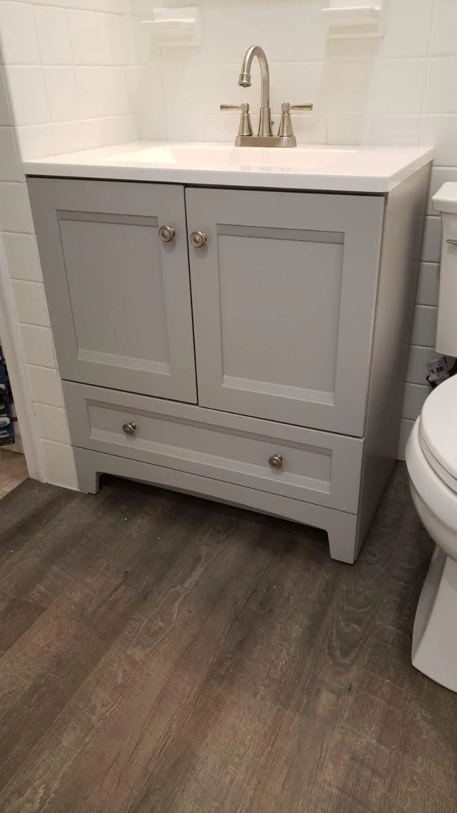 A bathroom vanity with a sink, faucet, and cabinet doors. The vanity is light gray with round knobs, situated next to a toilet, with wood-look flooring and white tiled walls.