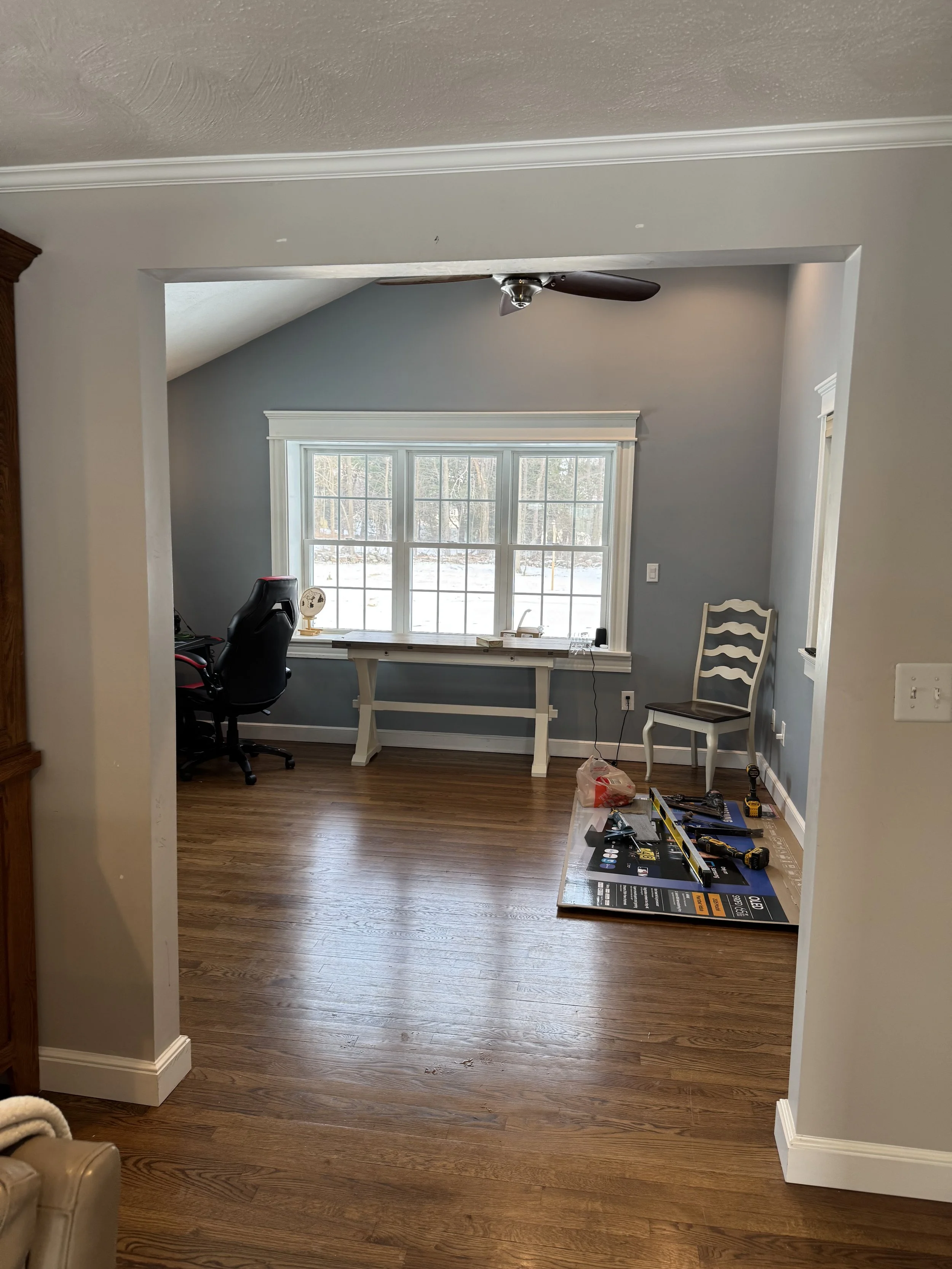 A room with light gray walls, a large window, and hardwood floors. There is a black office chair and a white table near the window, with some tools and a cardboard box on a mat on the floor. A white chair is in the corner and a ceiling fan hangs from