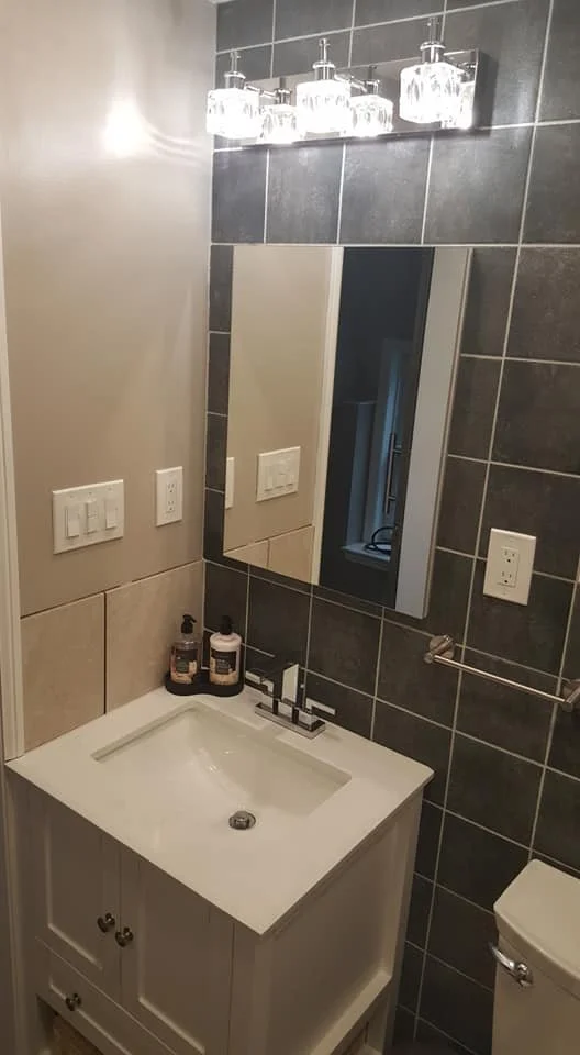 Bathroom with beige and dark gray tiled walls, a white vanity with a sink, soap and lotion bottles, a rectangular mirror, three light fixtures above the mirror, a towel bar, an electrical outlet, and a toilet.