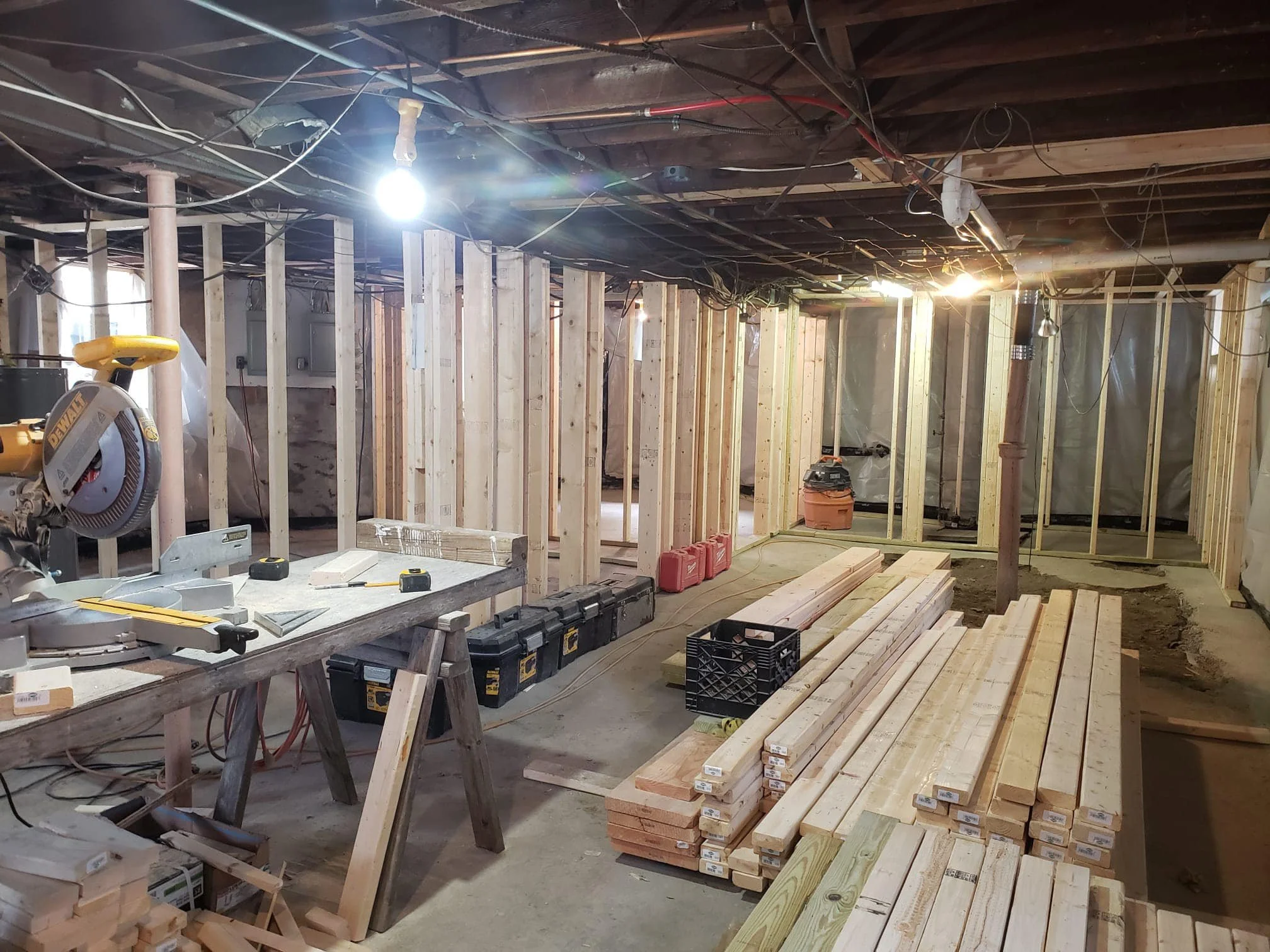 Interior view of a house under construction, with exposed wooden framing, construction tools, and materials like wood planks, a saw, and toolboxes.