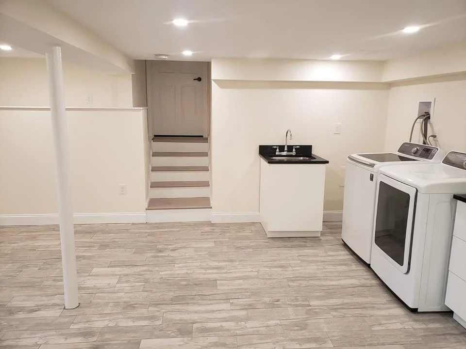 Basement laundry area with a washing machine, dryer, small black sink, and a staircase leading upstairs.