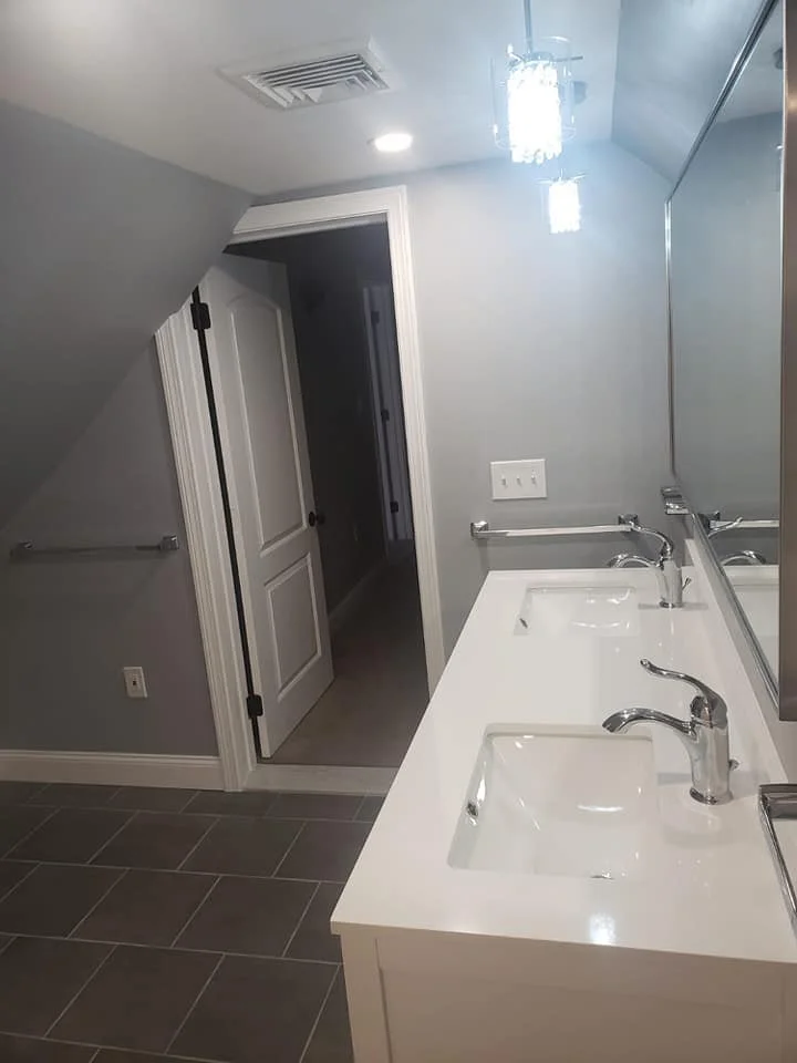 Bathroom with two sinks, large mirror, gray walls, tiled floor, ceiling light, and doorway leading to a darker room.