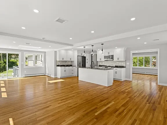 Empty modern open-concept kitchen with white cabinets, stainless steel appliances, island, and hardwood flooring.