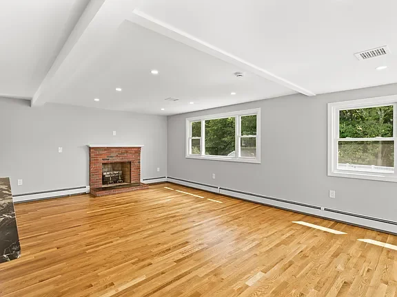 Empty living room with hardwood floors, gray walls, a brick fireplace, and multiple windows showing green trees outside.