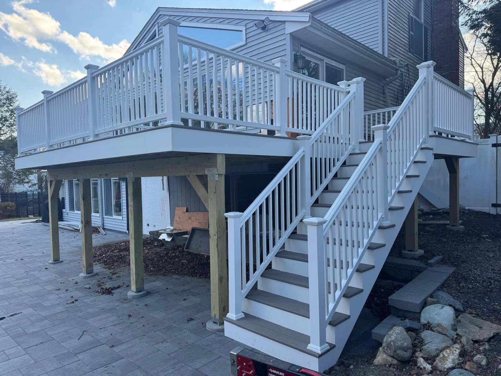 Newly built white wooden deck with stairs and railing attached to the back of a house, supported by wooden posts, with a backyard and a paved patio area below, under a partly cloudy sky.