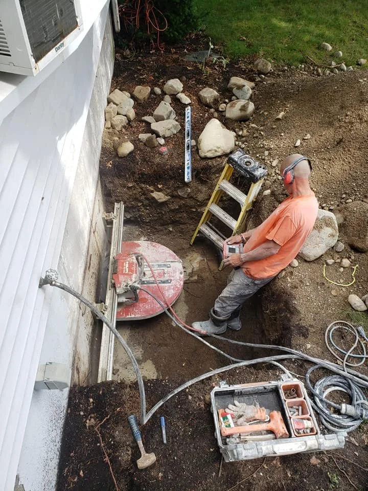 A worker wearing headphones and an orange shirt is operating a large circular saw at an outdoor construction site. The site includes a dug-out area with dirt and rocks, an extension ladder, and a wall of a building on the left side of the image.