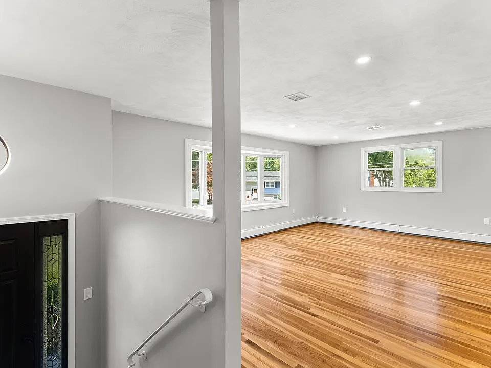Bright, empty living room with light gray walls, hardwood floors, three windows, and recessed ceiling lights.
