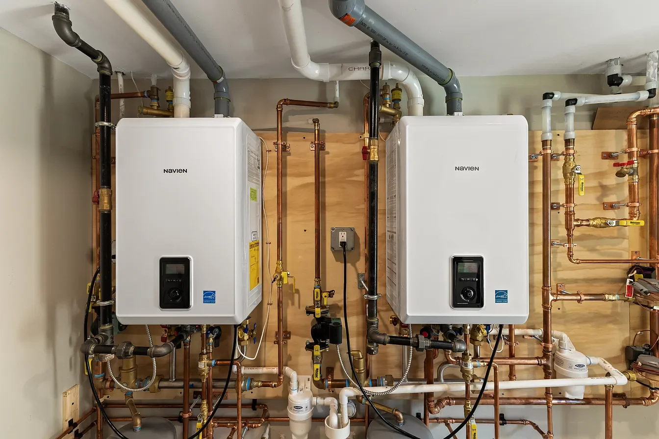 A utility room with two white Navien water heaters mounted on a wooden panel, surrounded by copper and plastic pipes, gauges, and valves.