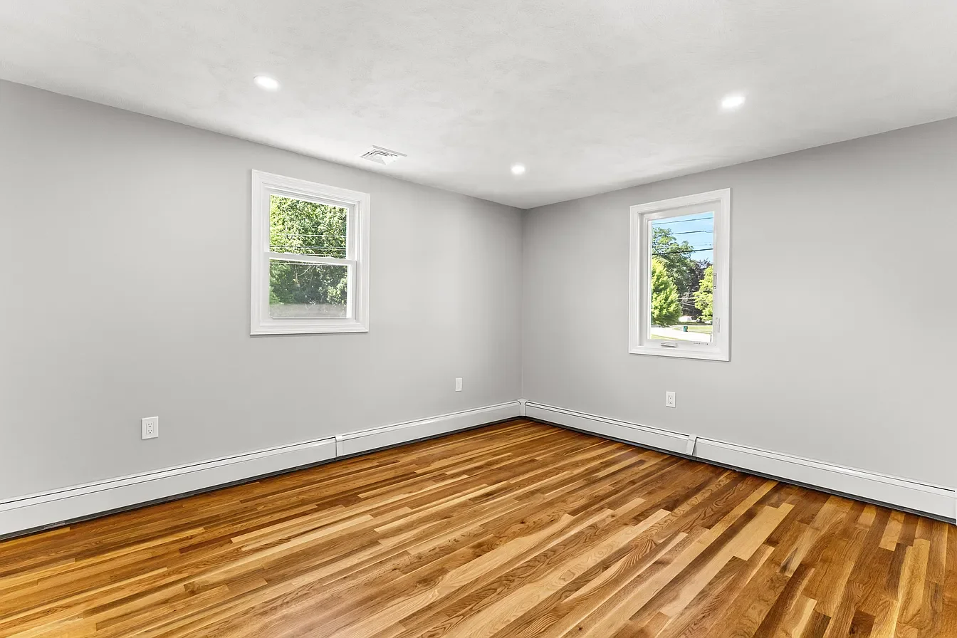 Empty room with hardwood floors, two windows, white walls, and recessed ceiling lights.