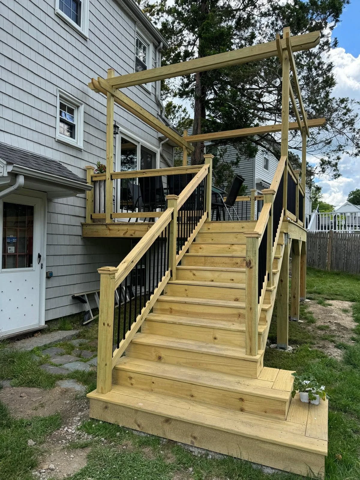 New wooden deck with stairs leading up to a back door of a house, featuring black metal railings and a dining table with chairs on the deck.
