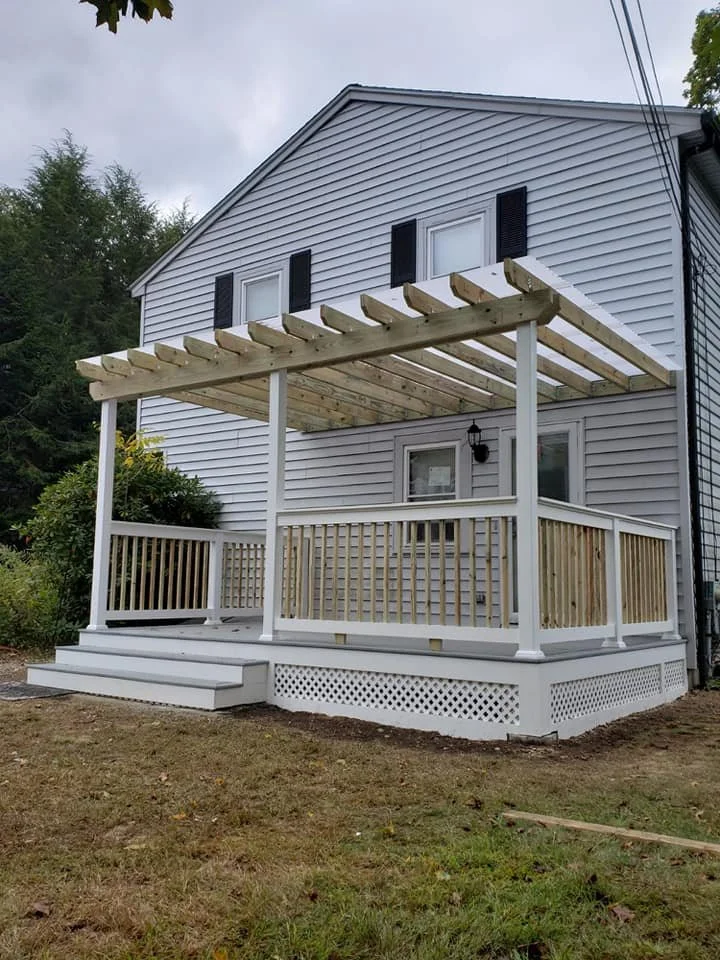 Front view of a newly built porch with stairs, railing, and partially constructed pergola on a white house.