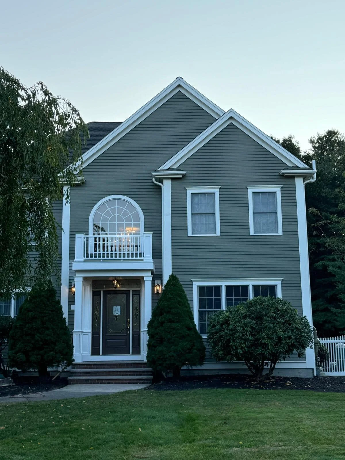 Front view of a two-story gray house with white trim and a black front door. The house has a small balcony above the entrance, framed by an arched window. There are neatly landscaped bushes and trees in the front yard, and a lawn leading up to the st