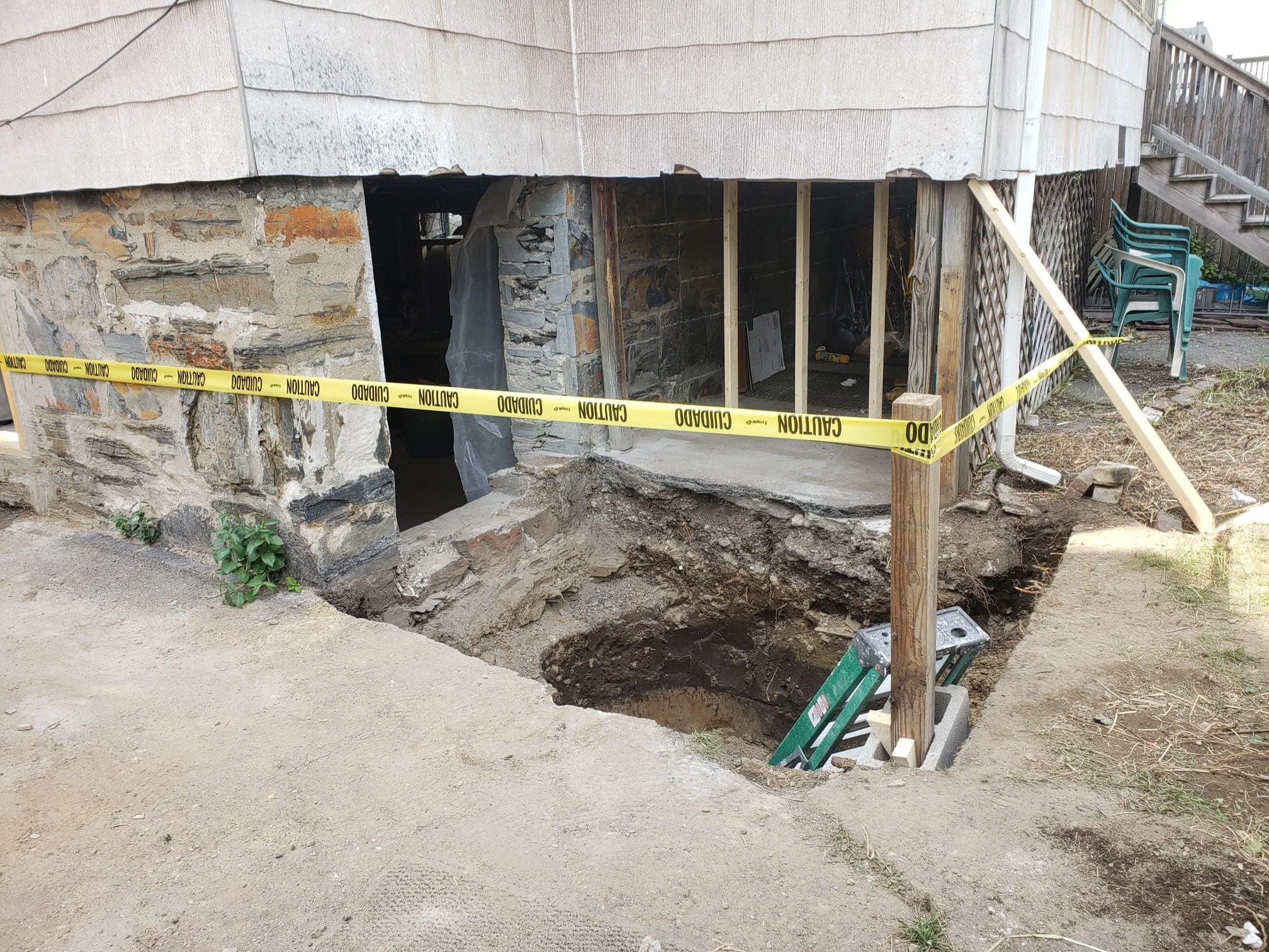 A house foundation is undergoing excavation and repairs, with a large hole near the corner of the house. The foundation walls are made of stone and brick, partially exposed, with wood supports and yellow caution tape around the work area. A green lad