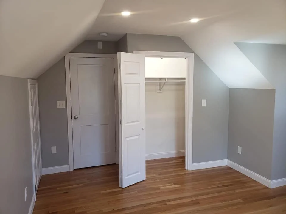 Empty room with light gray walls, hardwood floor, and a small closet with an open door. There is a closed door next to the closet, small slanted ceilings, and two ceiling lights.